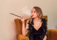 Élégante femme en robe de soirée noire vintage assise dans un fauteuil confortable et fumer la cigarette — Photo de stock