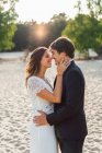 Люблячий чоловік обіймає красиву наречену в елегантній сукні і дивиться один на одного, стоячи на піщаному узбережжі під сонячним світлом — стокове фото