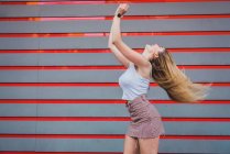 Молода жінка в повсякденному вбранні сміється і трясе волосся, стоячи на смугастій стіні — стокове фото