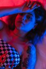 Молода жінка з рукою на лобі дивиться на камеру, лежачи в кімнаті з червоним і синім світлом — стокове фото