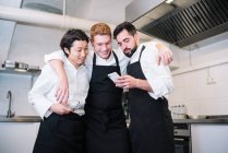 Три різних чоловіки в уніформі кухаря сміються і переглядають смартфони, стоячи на кухні ресторану разом — стокове фото