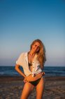 Расслабленная улыбающаяся женщина в бикини и рубашке, стоящая на пляже на закате — стоковое фото