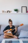 Mujer joven en camisa de gran tamaño a cuadros tocando la guitarra en el sofá - foto de stock