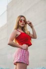 Attraente giovane donna in pantaloncini rossi e canotta toccare occhiali da sole e guardando la fotocamera mentre in piedi sulla strada — Foto stock