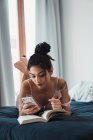 Гарненька брюнетка жінка розслабляється лежачи на ліжку з відкритою книгою і використовує смартфон — стокове фото