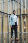 Calma elegante uomo etnico in piedi contro edificio di vetro e guardando la fotocamera — Foto stock