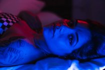 Junge Frau liegt auf Bett und blickt in Zimmer mit rotem und blauem Licht in die Kamera — Stockfoto
