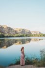 Woman in long summer dress standing on lake shore and looking over shoulder — Stock Photo