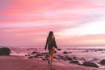 Visão traseira de ajuste sexy jovem fêmea em camisa preta andando na costa do oceano com areia e pedras ao pôr do sol com belo céu nublado rosa no fundo — Fotografia de Stock