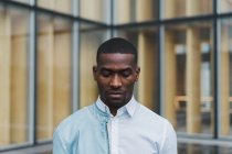 Ernster junger ethnischer Mann in weißem Hemd und heller Lederjacke auf einer Schulter, der gegen ein modernes Glasgebäude steht — Stockfoto