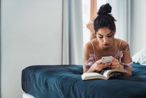 Гарненька брюнетка жінка розслабляється лежачи на ліжку з відкритою книгою і використовує смартфон — стокове фото