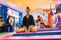 Junge Frau im karierten Oversize-Hemd blickt in die Kamera, während sie in der Halle mit verschiedenen Spielautomaten sitzt — Stockfoto