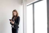 Элегантная женщина в костюме и с помощью планшета стоит в ярком дневном свете в современном офисе — стоковое фото