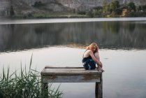 Чуттєва молода жінка сидить на пірсі на озері і обіймає коліна — стокове фото
