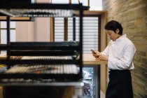 Side view of handsome Asian man in chef uniform standing in restaurant kitchen and browsing smartphone — Stock Photo