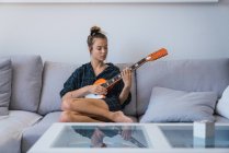 Jovem mulher em xadrez camisa oversize tocando guitarra no sofá — Fotografia de Stock