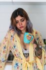 Schläfrige junge Frau im Seidenmantel mit einem Becher Heißgetränk, während sie morgens in der Küche steht — Stockfoto