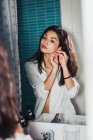 Selbstbewusste, elegante, brünette Frau im weißen Hemd, die Ohrringe vor dem Spiegel anlegt — Stockfoto