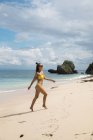 Счастливая женщина в жёлтом бикини гуляет по песчаному пляжу у океана — стоковое фото