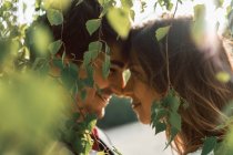 Vista lateral de la novia y el novio enamorados mirándose felizmente mientras están de pie en exuberante follaje verde a la luz del sol - foto de stock