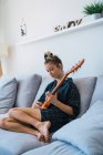 Young woman in checkered oversize shirt playing guitar on sofa — Stock Photo