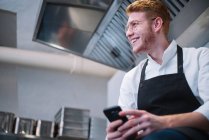 Вид сбоку на молодого человека в форме повара, опирающегося на кухонный прилавок и использующего современный смартфон, стоя на кухне ресторана — стоковое фото
