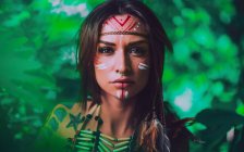 Attractive young woman with traditional Indian paintings on face looking at camera and standing in forest — Stock Photo