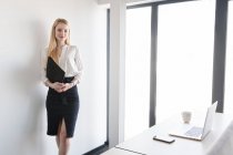 Элегантная женщина в костюме и с помощью планшета стоит в ярком дневном свете в современном офисе — стоковое фото
