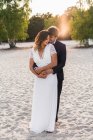 Liebender Mann umarmt schöne Braut in elegantem Kleid und schaut einander an, während sie an der Sandküste im Sonnenlicht steht — Stockfoto