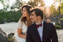 Щасливий елегантний чоловік і жінка у весільному вбранні, що обіймається пляжним каменем і дивиться усміхнено на сонячне світло — стокове фото
