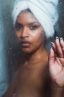 Чувственная афро-американка с полотенцем на голове, трогающая стакан в душе и смотрящая в камеру — стоковое фото