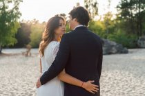 Loving man embracing beautiful bride in elegant dress and looking at each other while standing on sandy coast in sunlight — Stock Photo