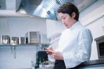 Vista lateral del hombre asiático guapo en uniforme de chef de pie en la cocina del restaurante y el teléfono inteligente de navegación - foto de stock