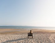 Вид ззаду жінка розслабляється на стільці на піщаному пляжі і дивиться на океан — стокове фото