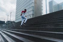Young fit ethnic man in sportswear running up stairs with glass modern buildings on background — Stock Photo