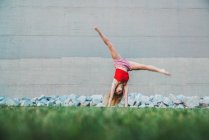 Young woman doing handstand on grass against concrete wall and stones — Stock Photo