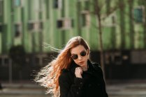 Stylish young redhead woman in sunglasses looking at camera on green background. — Stock Photo