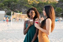 Giovani amiche che si rilassano sulla spiaggia con bevande in tazze e ridendo mentre chiacchierano al tramonto — Foto stock