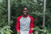 Lächelnder junger schwarzer Mann sitzt zwischen grünen Büschen im Gewächshaus und blickt in die Kamera — Stockfoto