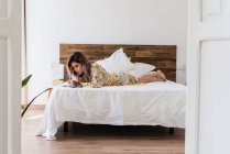 Jeune femme en robe de soie couchée sur le lit et faisant des croquis dans un bloc-notes dans une chambre élégante — Photo de stock