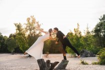 Seitenansicht von Mann und Frau im Brautkleid, die auf Baumstämmen über dem Sandstrand mit grünen Bäumen stehen und Händchen halten — Stockfoto