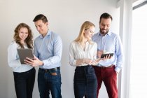 Кілька веселих елегантних чоловіків і жінок використовують різні гаджети, стоячи разом у світлому офісі — стокове фото
