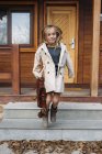 Happy blond girl in trench-coat with leather briefcase jumping in front of wooden house — Stock Photo