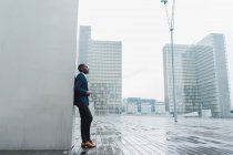 Afrikanisch-amerikanischer Geschäftsmann lehnt an einer Mauer im Freien mit modernen Gebäuden im Hintergrund — Stockfoto