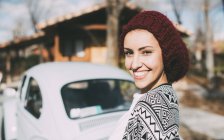 Молодая женщина в шерстяной шляпе стоит перед старой машиной и смотрит в камеру — стоковое фото