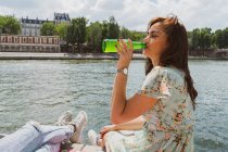 Молода жінка в літній сукні п'є воду на набережній — стокове фото
