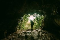 Mysterious view of traveler with backpack standing on rocky formation of cave entrance against tropical green forest. — Stock Photo
