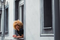 Молода етнічна жінка з афро волоссям, що спирається з вікна і дивиться в сторону денного світла — стокове фото