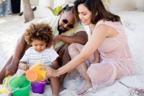Happy African-American parents and son sitting on sand and playing with buckets in sunny day on vacation — Stock Photo