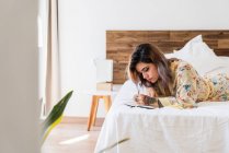 Jovem mulher em roupão de seda deitada na cama e fazendo esboços no bloco de notas no quarto elegante — Fotografia de Stock